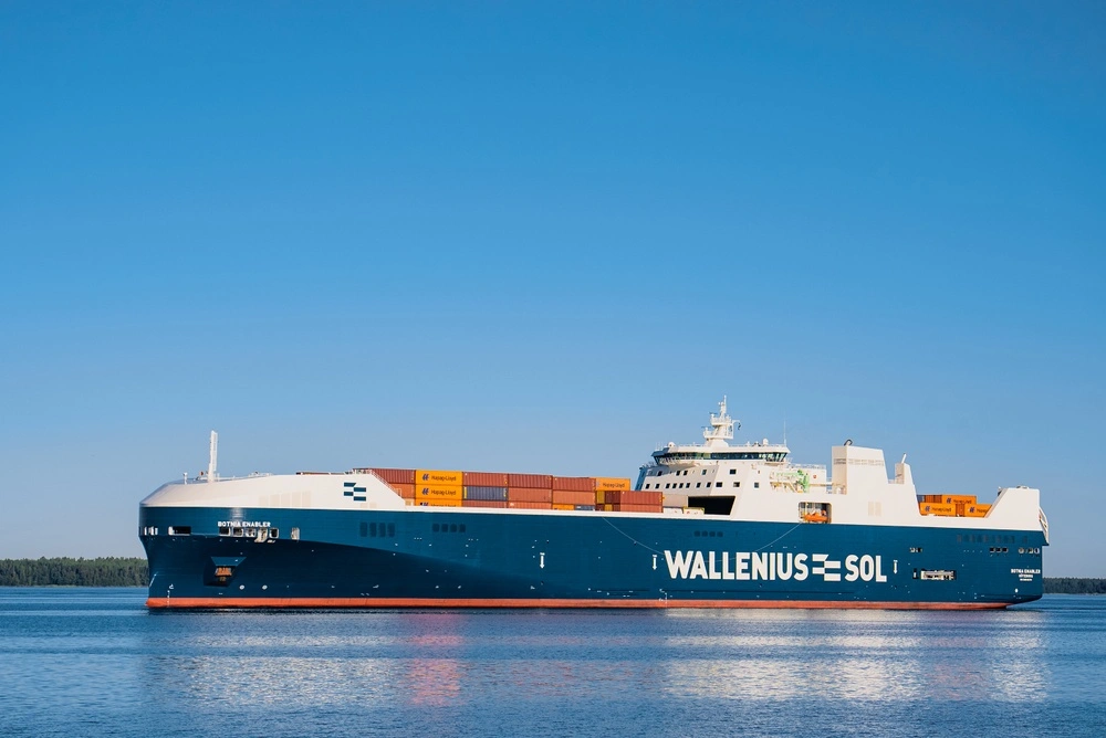 A large cargo ship with "WALLENIUS SOL" on its side sits on calm water under a blue sky. Several orange containers are stacked on the deck.