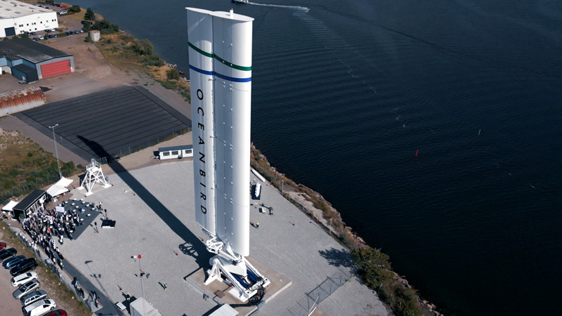A wing sail labeled "Oceanbird" sits on a launch platform next to water and a grassy area. Several people are gathered nearby.