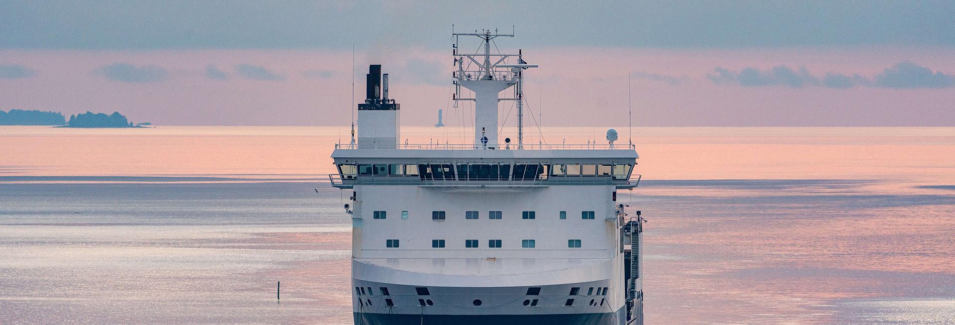 A large ferry boat on the water with a person standing on deck. Islands are visible in the distance under a pink sky.